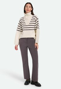 Maglione color crema chiaro con strisce grigie scure, dotato di un collo alto e texture a coste, abbinato a pantaloni a gamba larga grigi e scarpe nere.
