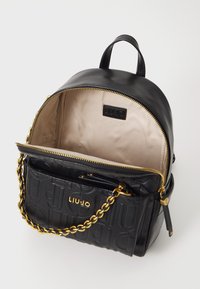 Zaino in pelle nera con forma arrotondata, accentato con catena dorata, logo stampato e interno in tessuto chiaro. Chiusura con zip e manico superiore.