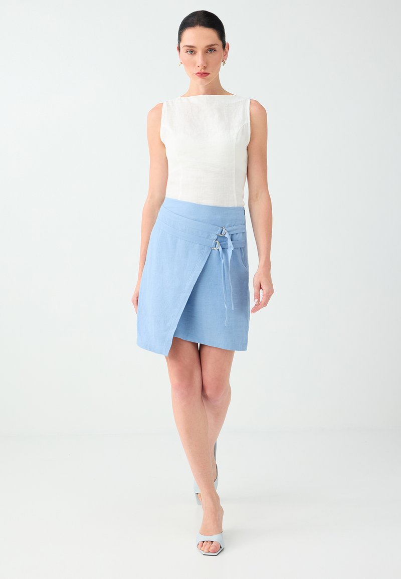 Femme aux cheveux foncés portant un haut blanc sans manches et une jupe portefeuille bleu clair, marchant vers l'avant sur un fond clair uni.