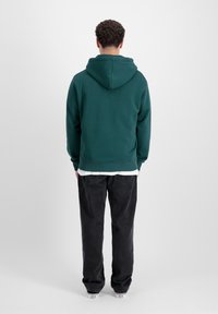 Felpa verde con cappuccio e cordino, tessuto morbido e polsini aderenti, abbinata a jeans neri e sneaker bianche. Vista posteriore su sfondo bianco.
