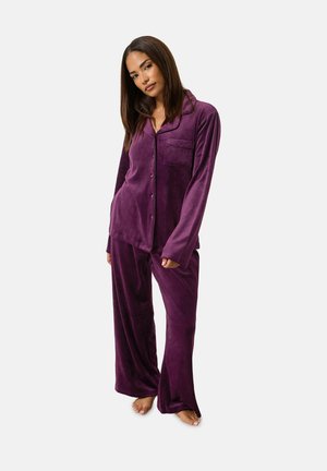 Femme debout pieds nus portant un pyjama en velours violet ample boutonné, à manches longues et pantalon à jambes larges.