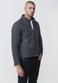 Férfi szürke farmerdzseki, állógallérral, cipzáras zsebekkel és vízszintes varrási részletekkel a vállakon. Jellemzője egy sima, kissé kopott textúra.