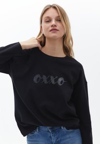 Czarna bluza o luźnym kroju, z błyszczącym szarym logo "oxxo" na piersi. Lekka tekstura z długimi rękawami.
