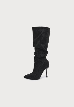 Botte noire souple montant jusqu’au genou, à bout pointu, à la texture finement ponctuée et au talon haut et fin, sur un fond clair.