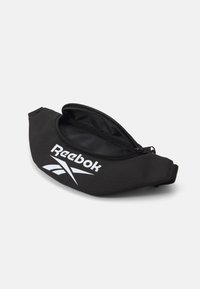 Čierna Reebok bedrová taška vyrobená z neoprénu s zaobleným dizajnom, ktorý má hladký povrch, logo značky a zipsové otváranie.