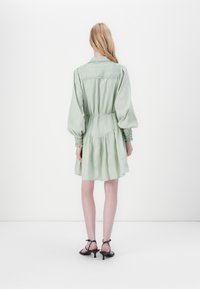 Hellgrünes, knielanges Kleid mit Puffärmeln, gerafften Akzenten an den Handgelenken und gerafftem Taillenbereich. Getragen mit schwarzen Riemchen-Heels.