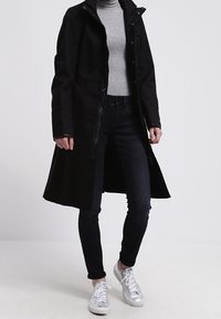 Abrigo negro largo con frente de botones, cuello de pie y bolsillos laterales, combinado con jeans oscuros y zapatillas plateadas. Tela texturizada visible.
