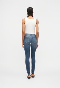 Άτομο με πλεξούδες, ντυμένο με λευκό αμάνικο τοπ, στενά μπλε τζιν και μαύρα σανδάλια με τακούνι, στέκεται μπροστά σε απλό φόντο.