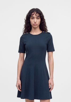 Une femme porte une robe bleu marine à manches courtes, ornée de boutons dorés aux épaules, sur un fond uni clair.