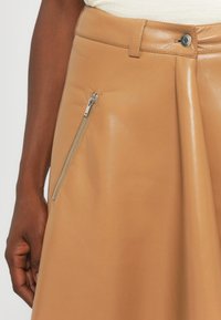 Suknja od smeđeg kože s glatkom teksturom, koja ima bočni džep s patentnim zatvaračem i pripijen struk s kopčanjem na dugme.
