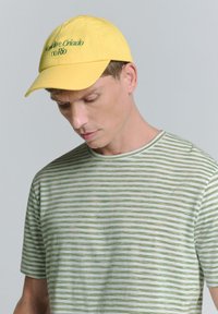 Geel baseballpet met groene geborduurde tekst, gecombineerd met een groen-wit gestreept shirt met korte mouwen van zachte stof.
