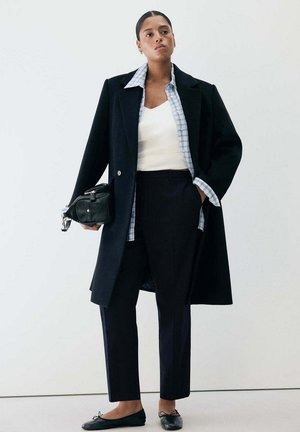 Femme portant un manteau noir, une chemise à carreaux, un haut blanc, un pantalon noir, des ballerines noires, tenant un sac noir, debout devant un fond blanc.