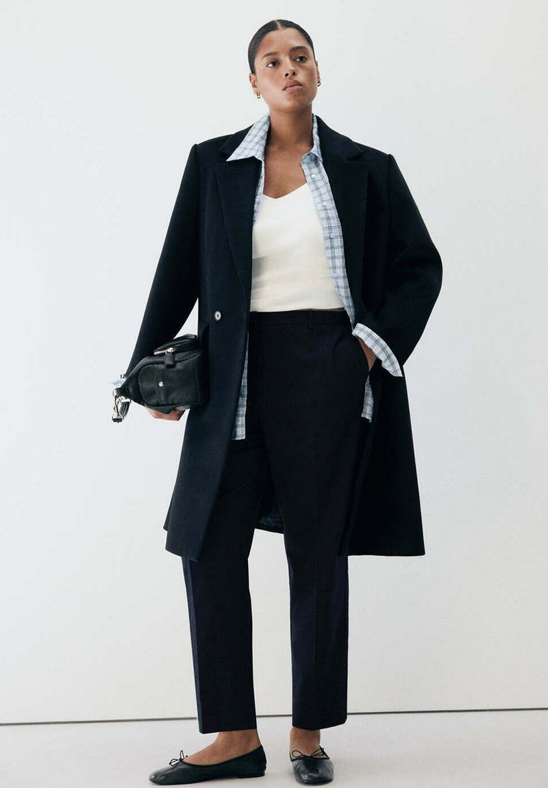Femme portant un manteau noir, une chemise à carreaux, un haut blanc, un pantalon noir, des ballerines noires, tenant un sac noir, debout devant un fond blanc.