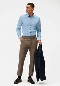 Hellblaues Hemd mit Knöpfen in Dunkel, kombiniert mit hellbraunen Hosen und einem dunkelblauen Blazer, der über dem Arm getragen wird. Schwarze Schuhe vervollständigen das Outfit.