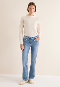 Beżowy sweter dzianinowy o prążkowanej fakturze w połączeniu z jasnoniebieskimi rozszerzanymi dżinsami. Modelka stoi z jedną ręką w kieszeni, ma na sobie białe trampki.