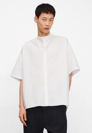 Uomo con indosso una camicia bianca a maniche corte oversize con collo alla coreana e pantaloni neri, in piedi su sfondo bianco uniforme.