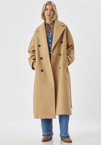 Camelfarvet dobbeltbrystet frakke i et blødt stof, med store klapper, sidelommer og sorte knapper, båret over denim og en mønstret bluse.