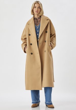 Kameelkleurige dubbelzijdige jas van een zachte stof, met grote revers, zijzakken en zwarte knopen, gedragen over denim en een gebloemd blouse.