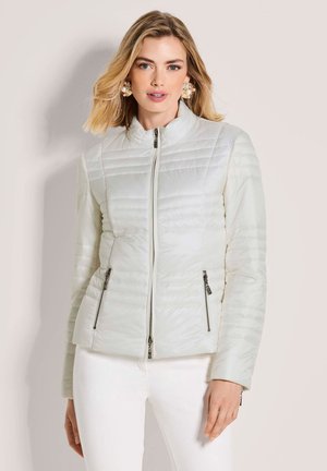 Frau trägt eine weiße, gestepptet Reißverschlussjacke mit Reißverschlusstaschen, kombiniert mit weißer Hose und großen runden Perlenohrringen.