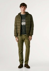 Kaki puffjacka med huva, bärs över en mörk tröja med vita och orange mönster, kombinerat med olivgröna byxor och bruna stövlar.