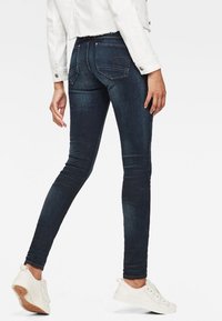 Persona vestida con jeans ajustados azul oscuro, zapatillas blancas y una chaqueta blanca corta, de pie con un pie ligeramente elevado.