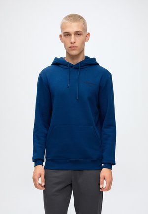 Jeune homme aux cheveux blonds courts portant un sweat à capuche bleu et un pantalon gris foncé, debout sur un fond blanc uni.