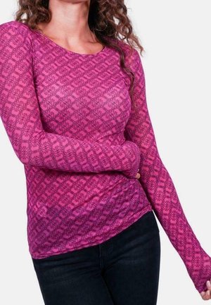 Vrouw draagt een roze top met lange mouwen en patroon en donkere broek, poseert met één arm gebogen over haar taille tegen een effen achtergrond.