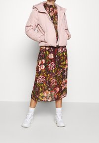 Růžová vatovaná bunda s kapucí a elastickými manžetami přes květované šaty po kolena v tmavě červené, zelené a růžové barvě, doplněné bílými teniskami.