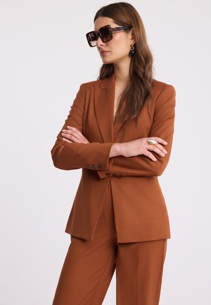 Kobieta z długimi brązowymi włosami, nosząca okulary przeciwsłoneczne z tortoise shell, rdzawosutannowy garnitur, dużą pierścionek i złote kolczyki, stoi z założonymi rękami.