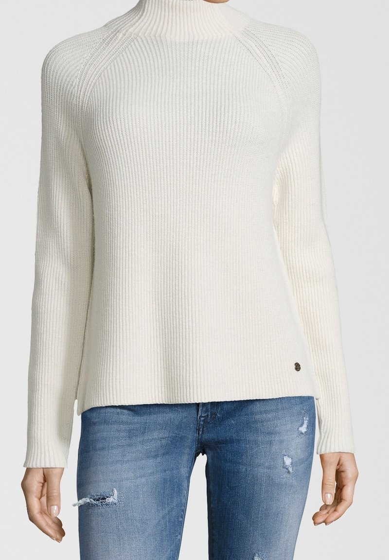 Femme portant un pull à col roulé côtelé crème et un jean bleu déchiré, debout, les bras détendus le long du corps.