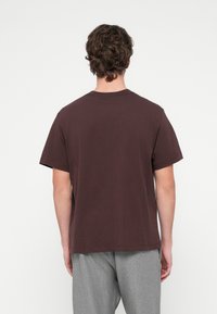 Καφέ βαμβακερό μπλουζάκι με κοντά μανίκια, στρογγυλή λαιμόκοψη και άνετη γραμμή. Το ύφασμα φαίνεται απαλό και λεία.