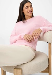 Hellrosa, gestrickter Pullover mit lockerer Passform, langen Ärmeln und rundem Halsausschnitt. Beigefarbene Hose, sitzt auf einem runden, gepolsterten Stuhl.