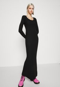 Vestido maxi preto canelado com mangas longas e decote em forma de barco, combinado com botas de atacadores rosa brilhante, criando um visual de alto contraste.