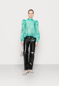 Blusa verde menta translúcida com mangas bufantes, combinada com calças pretas de brilho intenso. A modelo segura uma pequena bolsa xadrez e usa saltos altos.