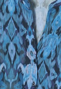 Blusa de estampado azul que presenta una mezcla de tonos azules claros y oscuros, fabricada en tejido ligero y con un cierre frontal abotonado con botones oscuros.