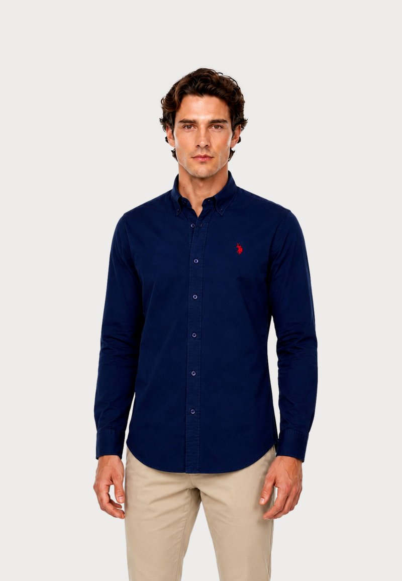 Uomo che indossa una camicia blu navy con bottoni e un logo rosso sul petto e pantaloni beige, in piedi davanti a uno sfondo chiaro e semplice.