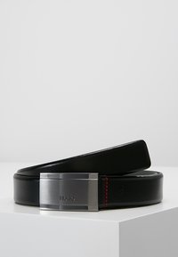 Ceinture en cuir noir avec une boucle rectangulaire en métal argenté, présentant un logo en relief et des accents de couture rouge. Texture lisse et design épuré.