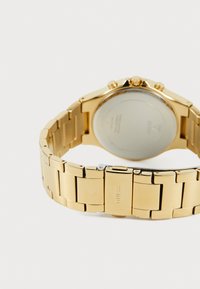 Montre-bracelet en métal doré avec une finition polie, un boîtier rond et une couronne. Présente un dos lisse et réfléchissant, ainsi que des détails de fermoir.