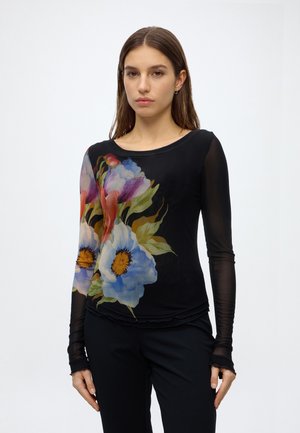 Femme portant un haut noir à manches longues avec un grand imprimé floral coloré sur le devant et des manches transparentes, se tenant devant un fond blanc uni.