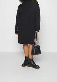 Černé oversized pletené šaty s žlutými bočními akcenty, černé šněrovací stylové boty a černá kabelka s řetízkem.