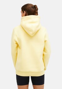 Gelber Hoodie mit großer Kapuze, gerippten Bündchen und Saum sowie weichem Stoff. Ansicht von hinten, kombiniert mit schwarzen Shorts.