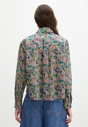 Frau mit langen braunen Haaren, die ein rosa geblümtes Langarmshirt und blaue Jeans aus Denim trägt, von hinten vor einem einfarbigen weißen Hintergrund gezeigt.