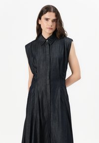 Mørk denim ermeløs skjortekjole med knappelukking foran, spiss krage og plissert skjørt. Glatt tekstur, minimalistisk design.