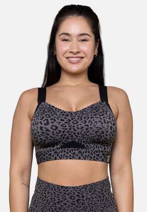 Smiling woman with long dark hair wearing a gray and black leopard print sports bra and matching high-waisted leggings against a white background.