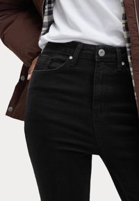 Svarta denimjeans med fem fickor, kraftig sömnad, knäppning med knapp och silverdetaljer. Bärs med en vit skjorta och brun jacka.