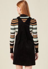 Mustapintainen corduroy-mekko, jossa on säädettävät olkaimet, tiukka yläosa ja levenevä hame, kerroksittain värikkään raidallisen pitkähihasen topin päällä.