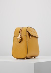 Gul lædertaske med en struktureret overflade, guld lynlås og sorte detaljer. Firkantet design med klap foran og hardwaredetaljer på siden.