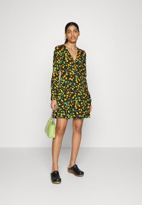Vestido preto com padrão de frutas amarelas e verdes, mangas longas, decote em V, com bainha em babados, combinado com uma bolsa verde e tamancos de madeira pretos.