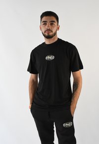 Camiseta de algodón negra con un escote redondo y mangas cortas, con un logo en el centro. Combinada con pantalones de chándal negros a juego con acentos de logo.