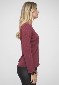 Langärmeliges, dunkelrotes Oberteil mit rundem Ausschnitt und schwarzen Spitzenakzenten an den Bündchen, kombiniert mit schwarzen Leggings. Glattes, strukturiertes Material.